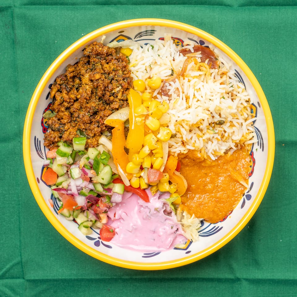 Overhead view of a colorful bowl with rice, curried meat, salad, corn, peppers, and pink sauce.