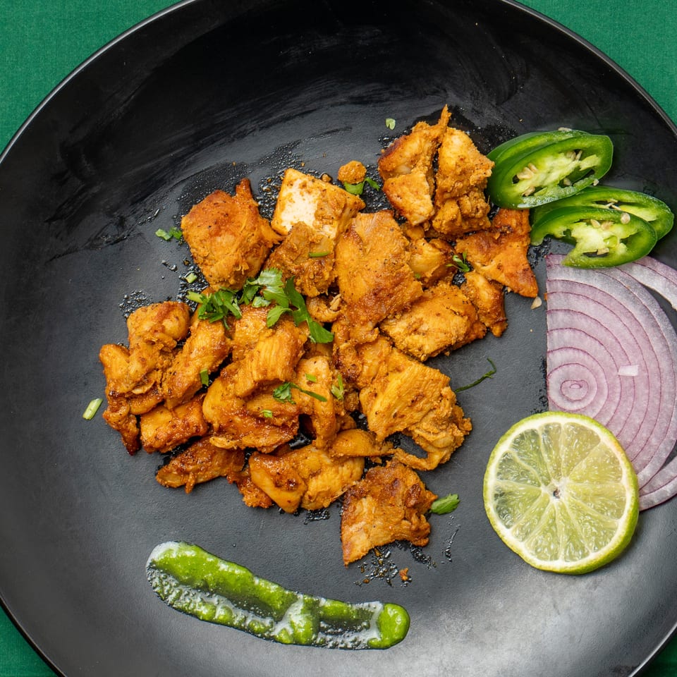 Spicy chicken tikka pieces on a black plate with lime, red onion, jalapeños, and green sauce.