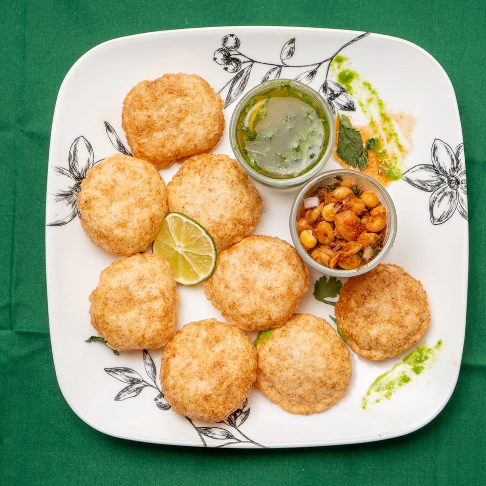 A white plate with nine golden-brown pani puri, two bowls of filling, and a lime wedge.
