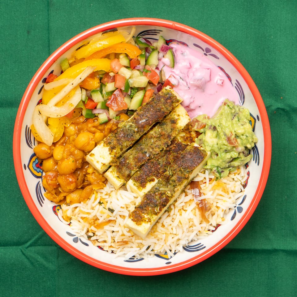 Colorful bowl with rice, paneer, chickpeas, guacamole, pink yogurt, and vegetable salad.