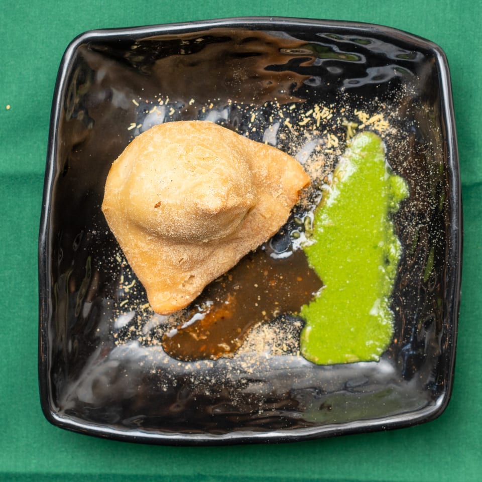 A golden-brown samosa on a black plate with green and brown sauces, sprinkled with spices.