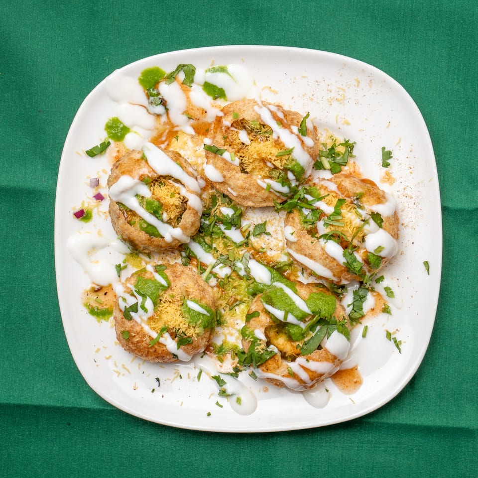 A white plate of dahi puri, topped with yogurt, green chutney, and cilantro, on a green tablecloth.