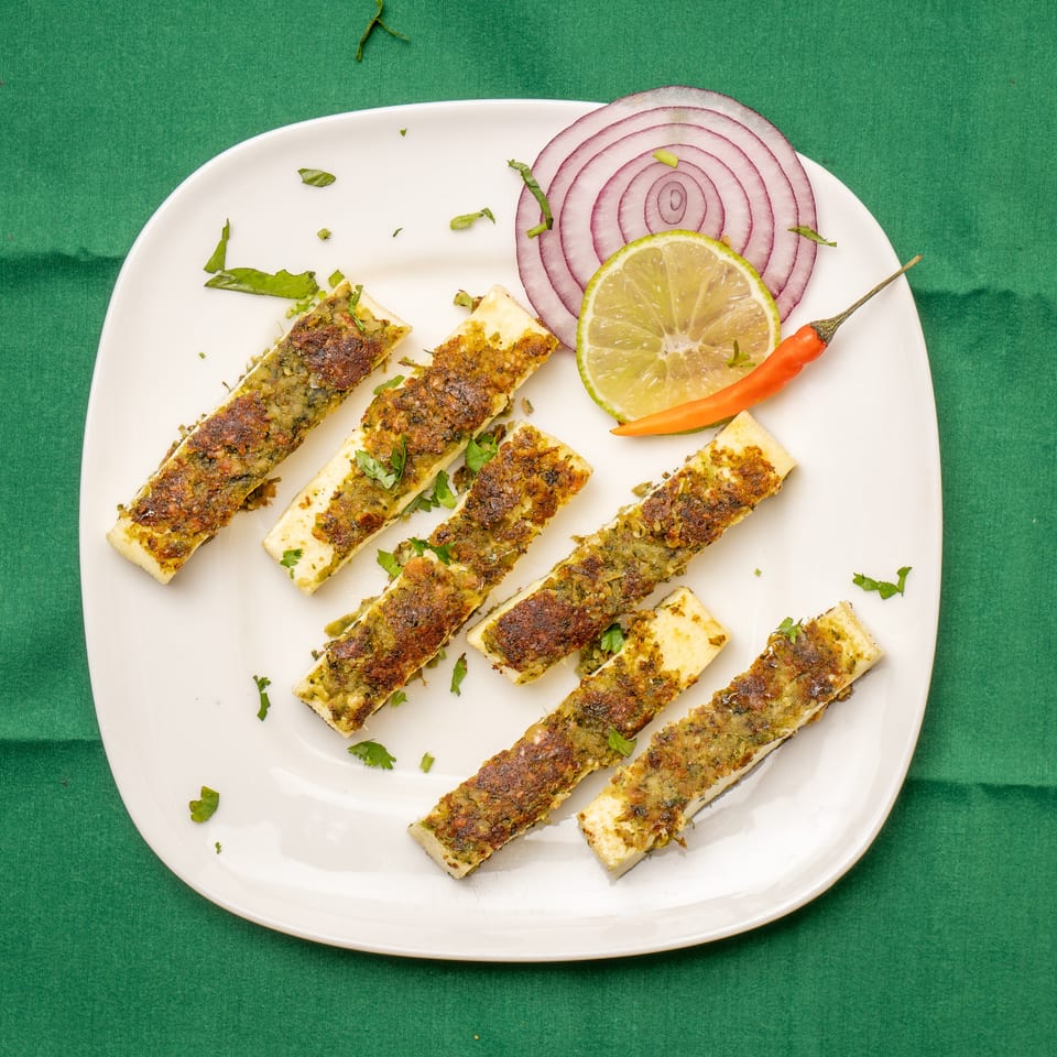 Grilled paneer sticks with green herb crust, garnished with red onion, lime, and chili on a white plate.
