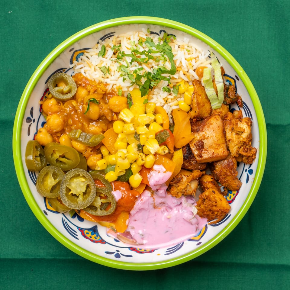 Colorful bowl with rice, chickpeas, corn, peppers, chicken, jalapenos, and pink sauce on green cloth.
